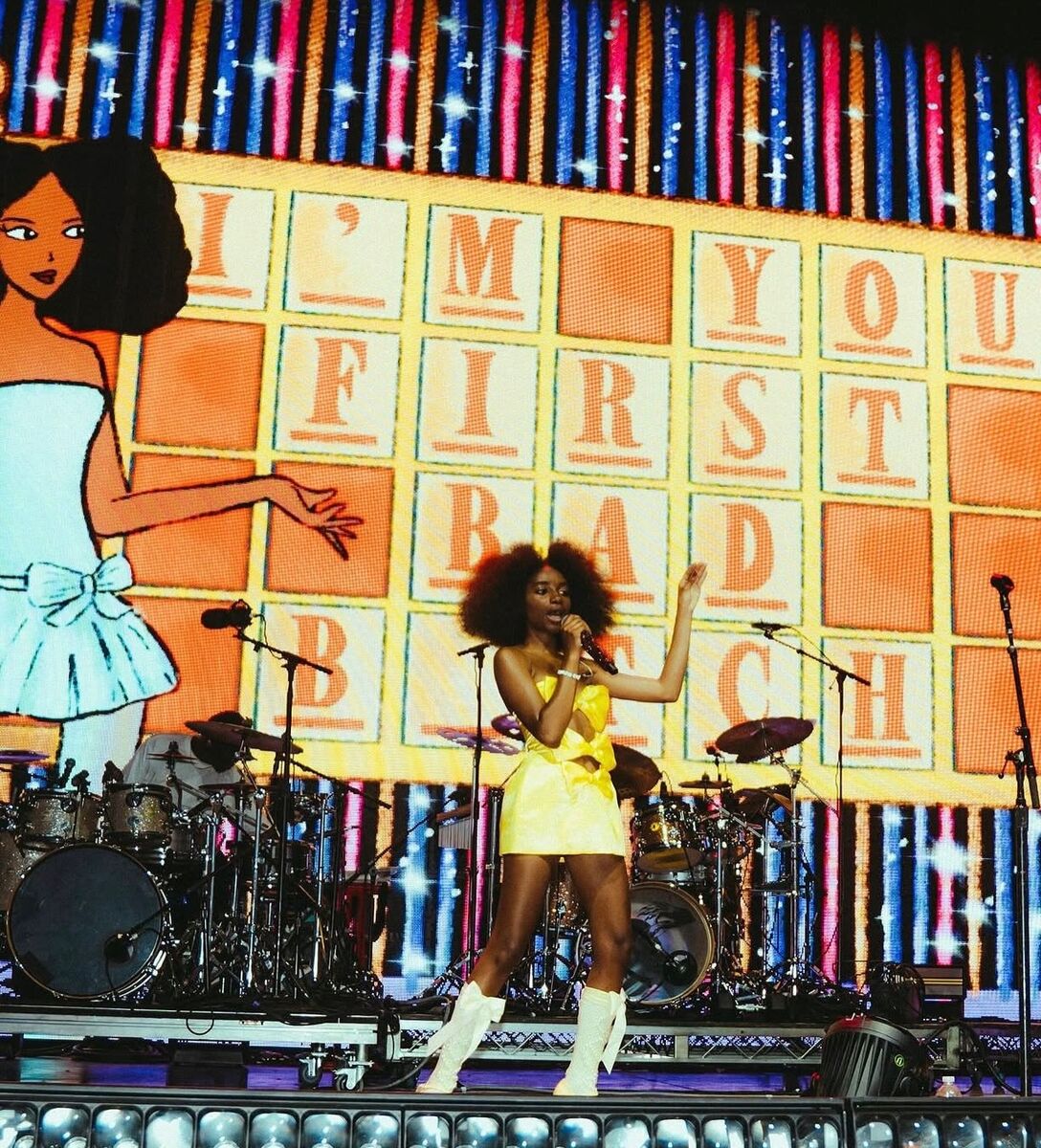 Performer in a yellow costume singing on stage in front of a large painted set